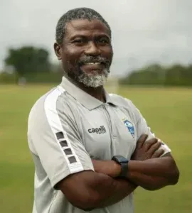 Man with crossed arms, wearing a grey polo shirt with a logo, smiling outdoors on a green field.