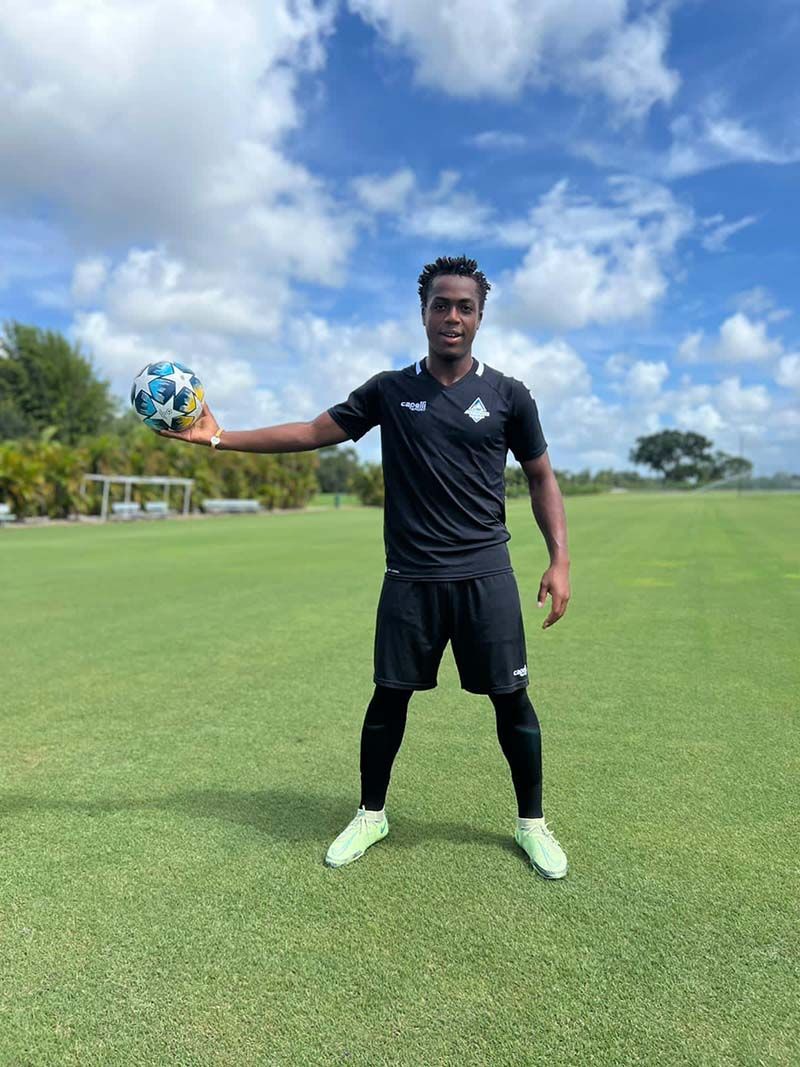 Soccer player in black uniform holding a soccer ball on a green field under a blue sky.