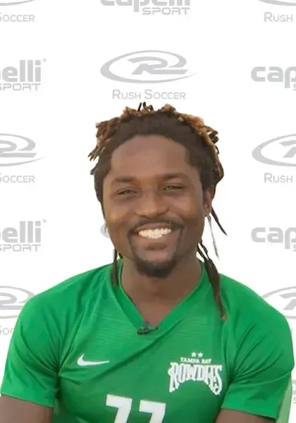 Lucky Mkosana green soccer jersey with dreadlocks, smiling, near a logo.