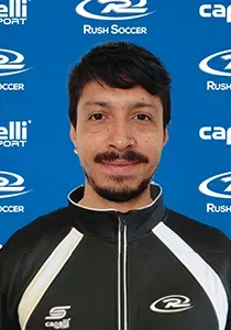 Man with short dark hair and a mustache wearing a black and white jacket, smiling at the camera. Soccer logo background.