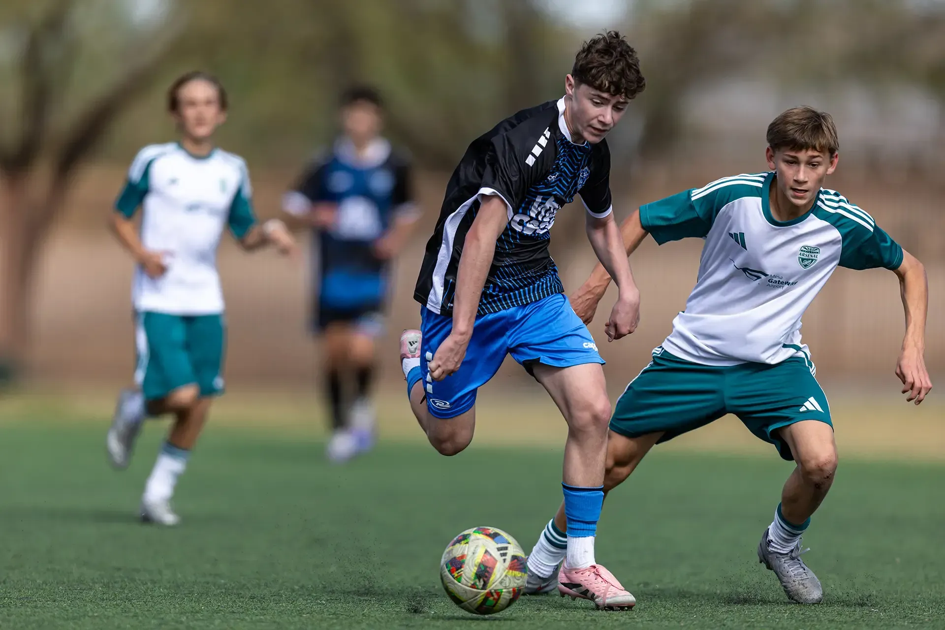 Soccer game on green field. Players in light blue and dark blue uniforms. Referee watches. Mountains in background.