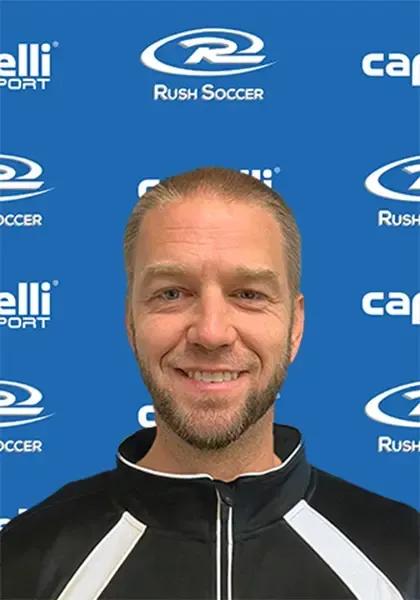 Man with a short beard smiling, wearing a black and white jacket, against a blue background with soccer logos.