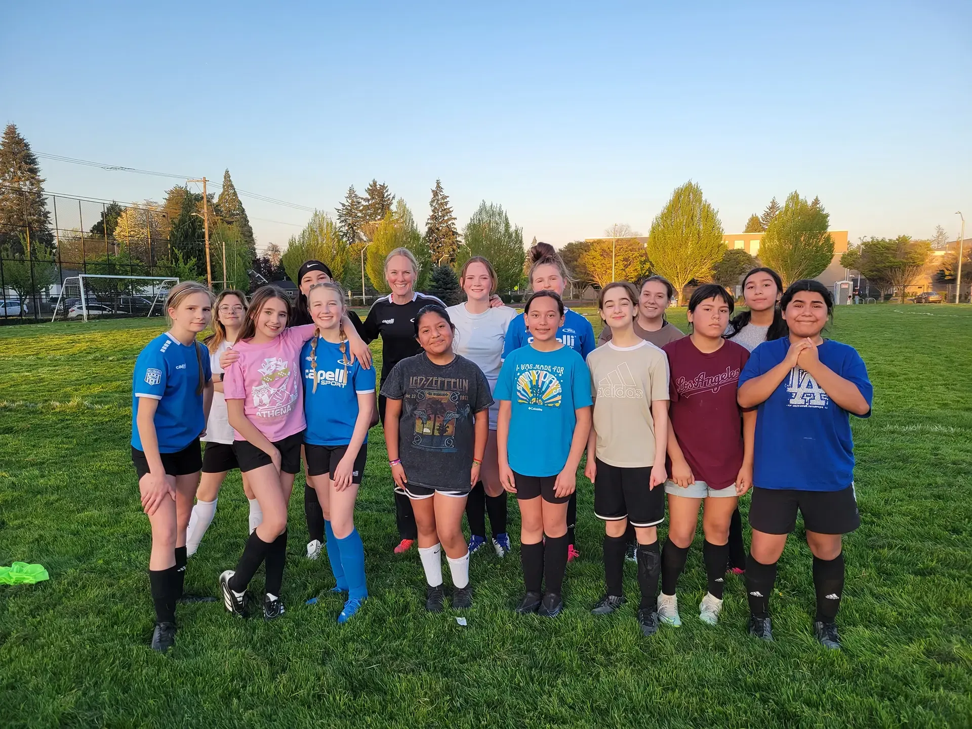 Soccer team poses on a grassy field in front of trees. Several girls wear soccer gear and smile.