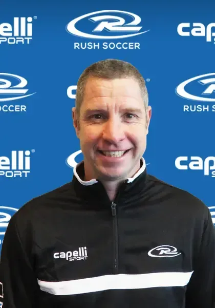 Man smiling in a black jacket with soccer logos, against a blue backdrop.