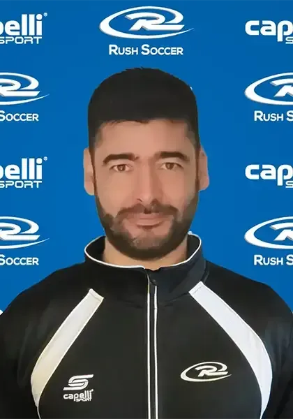 Man with a beard in a black and white jacket, in front of a blue background with soccer logos.