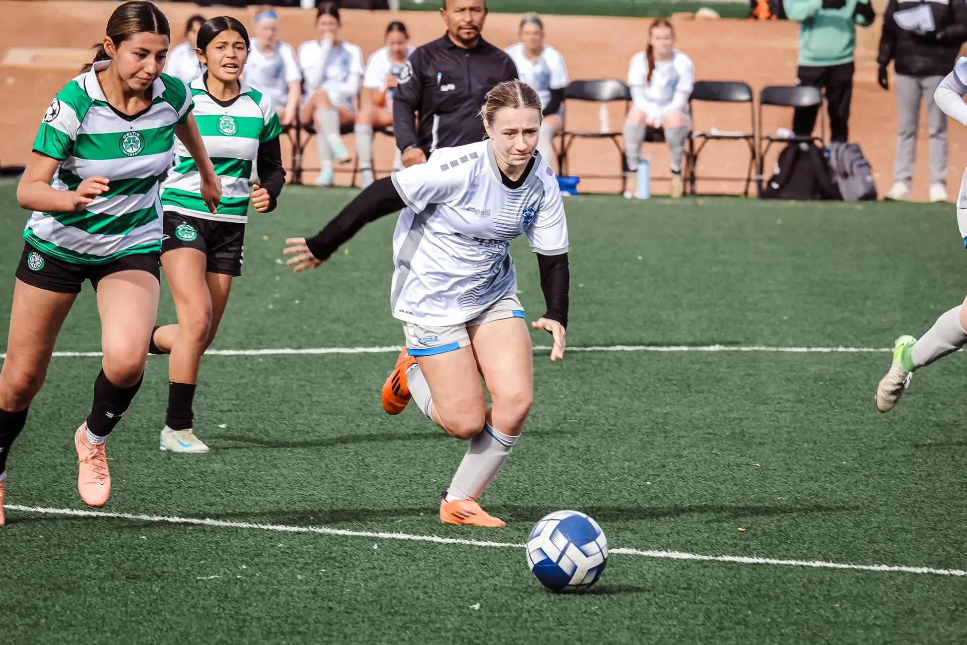 Soccer game on green field. Players in light blue and dark blue uniforms. Referee watches. Mountains in background.