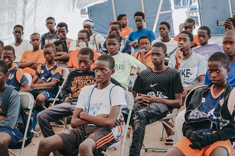 A group of young people seated outdoors, some wearing athletic shirts, appearing attentive.