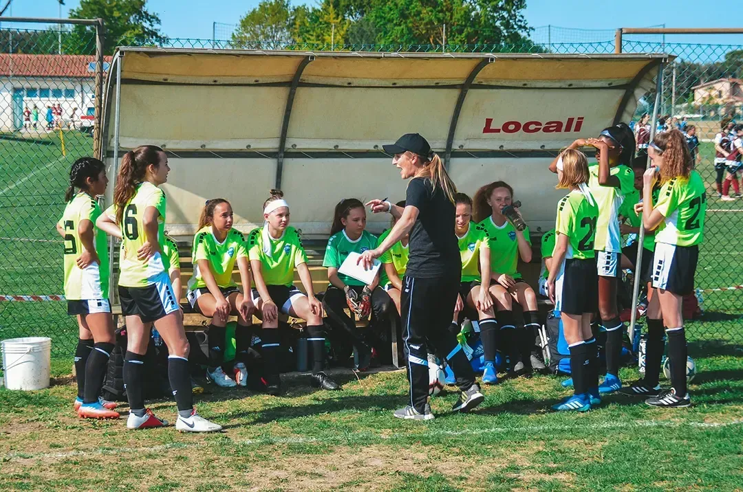 A coach talks to a team of young players on the bench during a soccer game. They're wearing green and black uniforms.