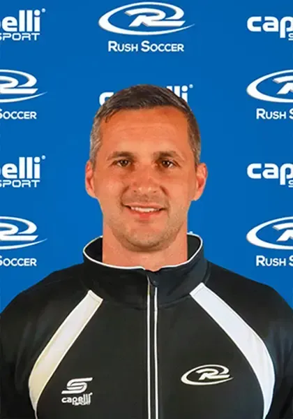 Soccer coach in black and white jacket, smiling, in front of a blue banner with 