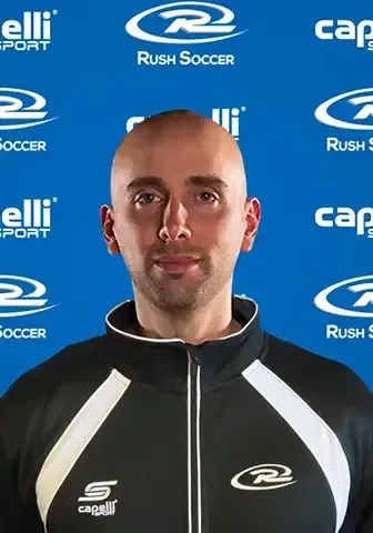 Bald man in black and white jacket, standing in front of a blue backdrop with soccer-related logos.