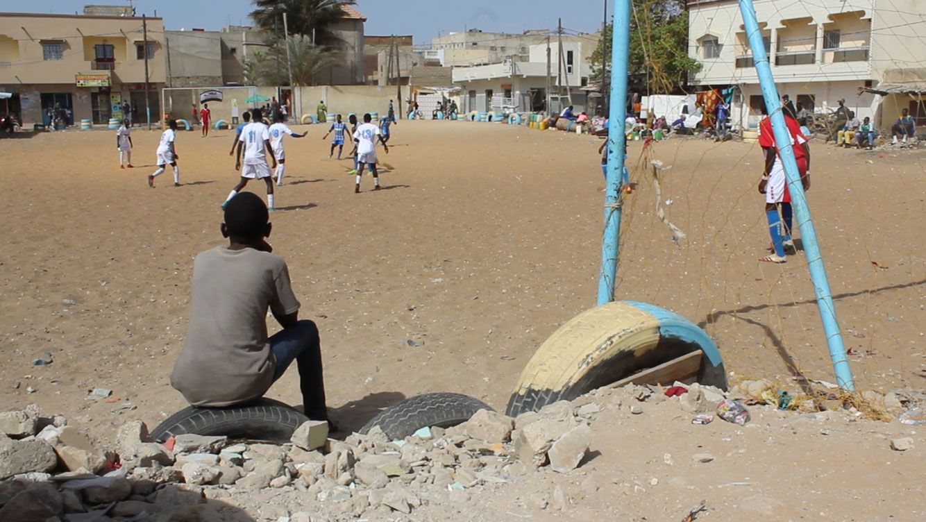 A person watches a soccer game on a dirt field. Players in white and blue jerseys are running. Buildings in background.