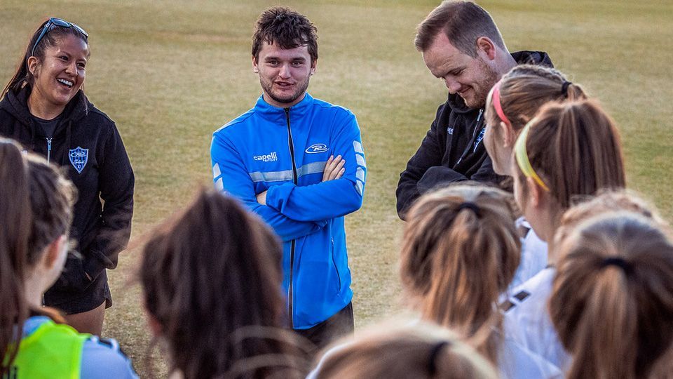 A group of people on a field, including a man in blue and women in sportswear, appear to be in discussion.