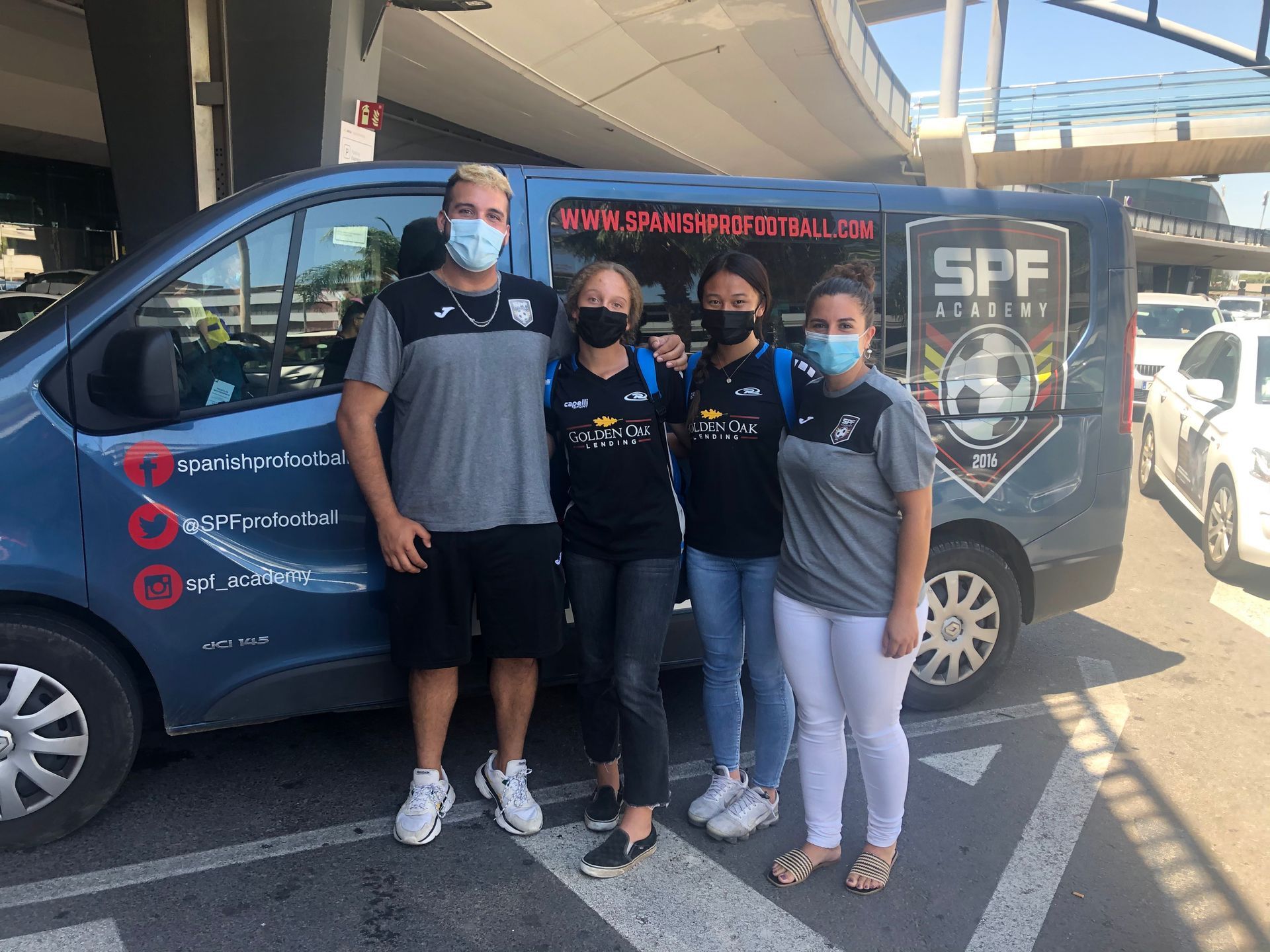 Four people pose beside a blue van with logos. They are wearing masks. The van is in front of a building in bright sunlight.