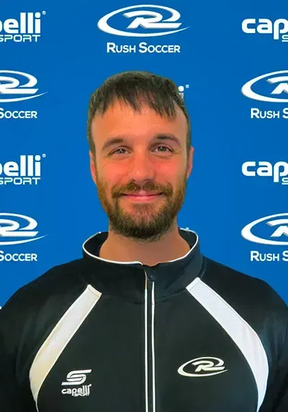 Man with beard wearing a black and white athletic jacket in front of a blue background with soccer logos.