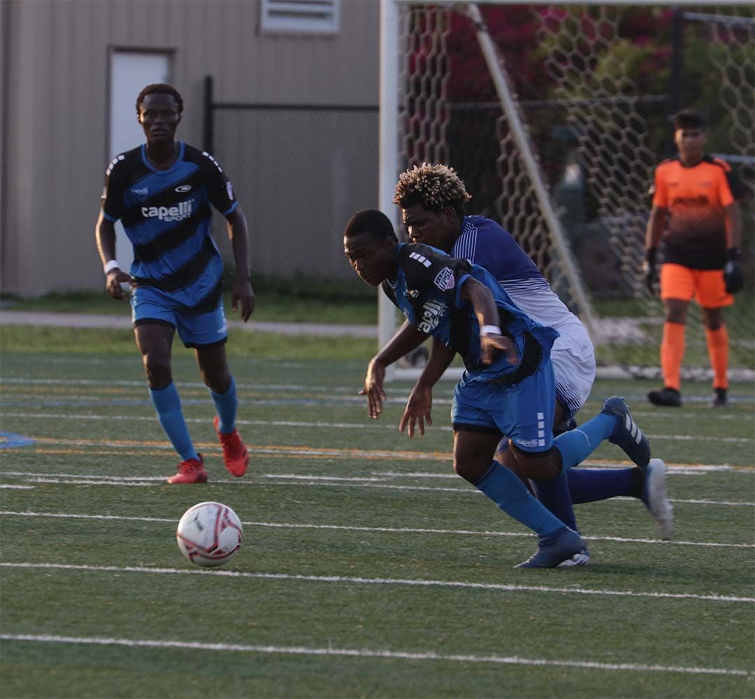 Soccer players on a green field. One in blue tries to kick the ball while another in blue/purple attempts to defend. 