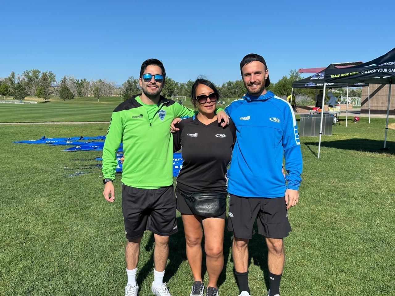 Three people posing on a grassy field; two men in athletic wear, one in a black outfit, and a woman in sunglasses. Sunny day.