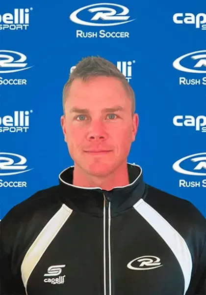 Man in a black and white sports jacket against a blue background with soccer logos.