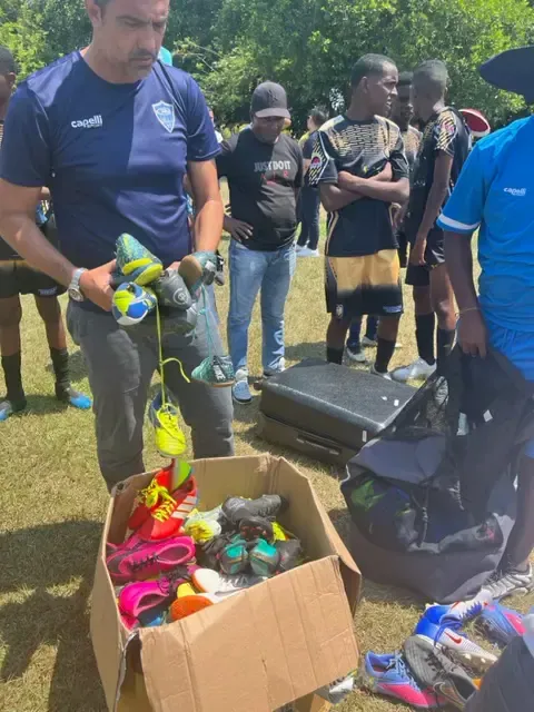 Man sorting through a box of athletic shoes outdoors, possibly for a team. Other people watch.