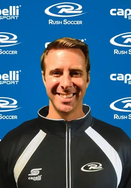 Man smiling, wearing a black and white athletic jacket. Blue background with soccer team logos.