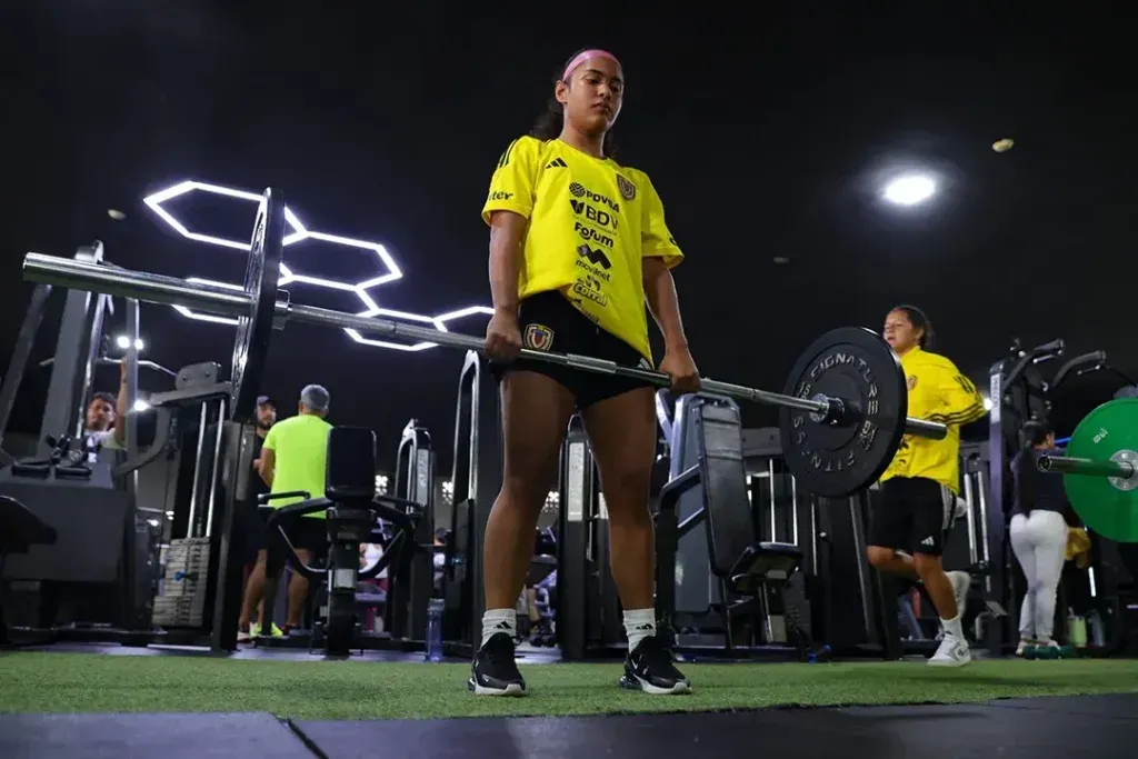 Woman lifting weights with barbell in a gym, wearing yellow shirt and black shorts.