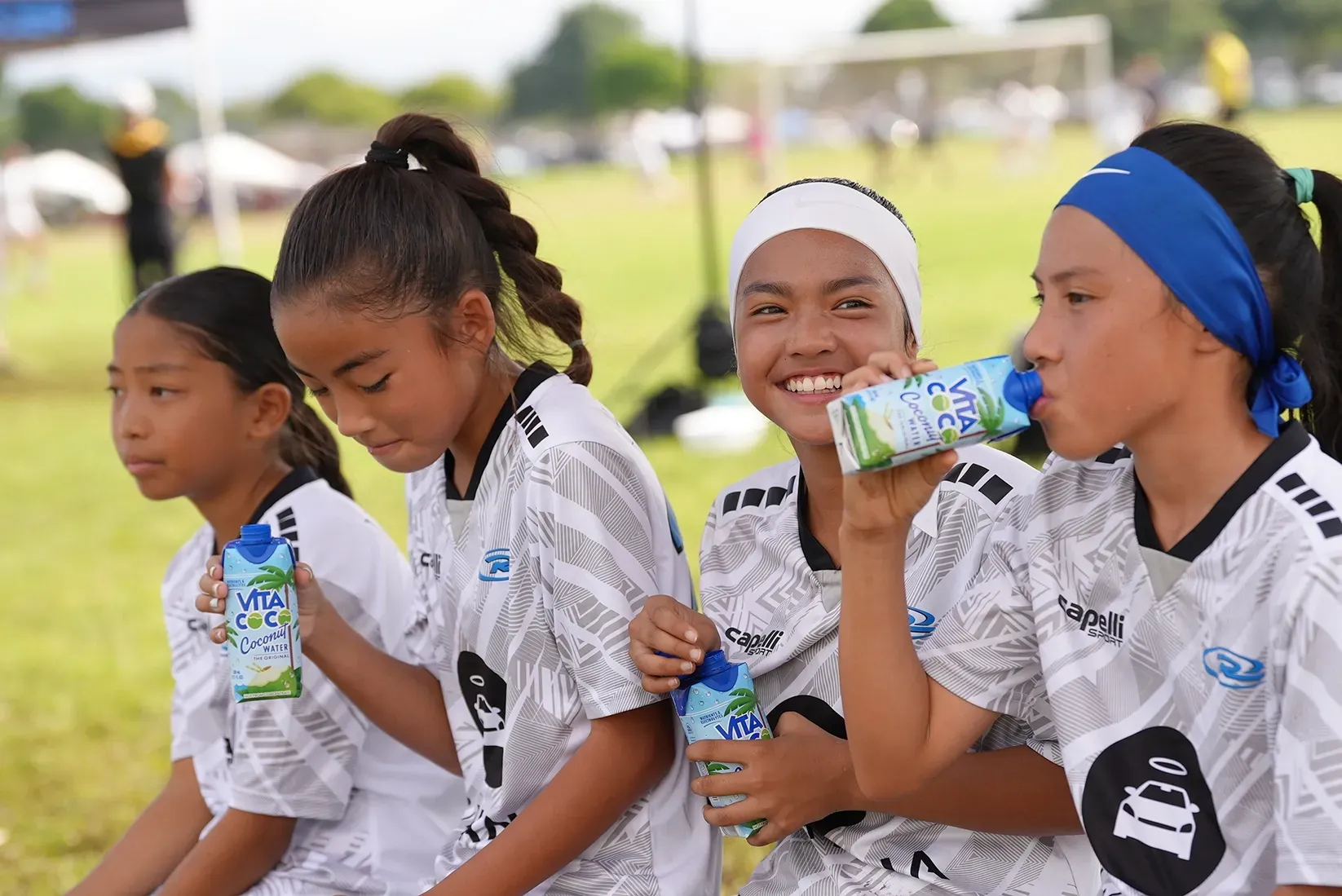 Four young soccer players in white jerseys rest on a bench, drinking from blue cartons.