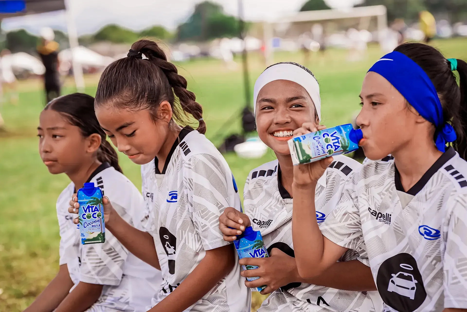Four soccer players in jerseys drink beverages on a grassy field. One smiles, others look on.
