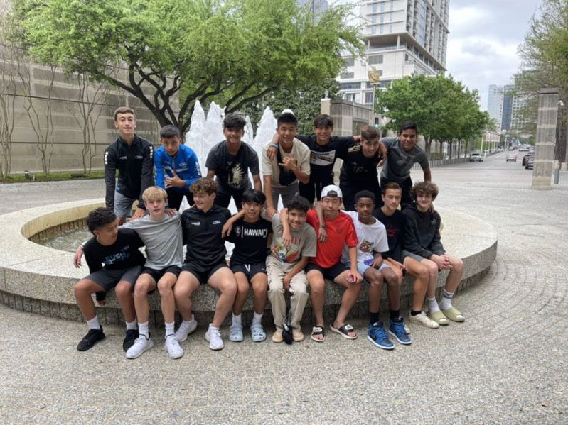 Group of boys posing in front of a fountain. They are sitting and standing, smiling. Buildings and trees in the background.