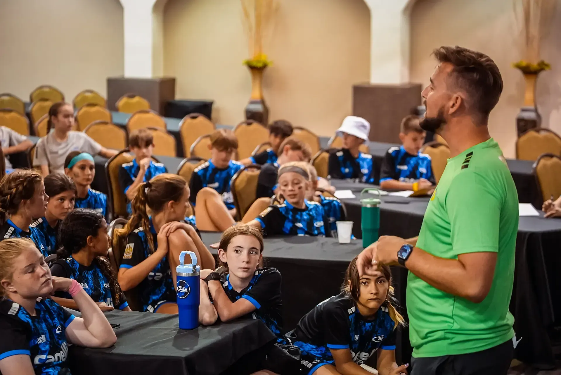 Soccer coach in green shirt speaking to team of young players in a meeting room; team listening.
