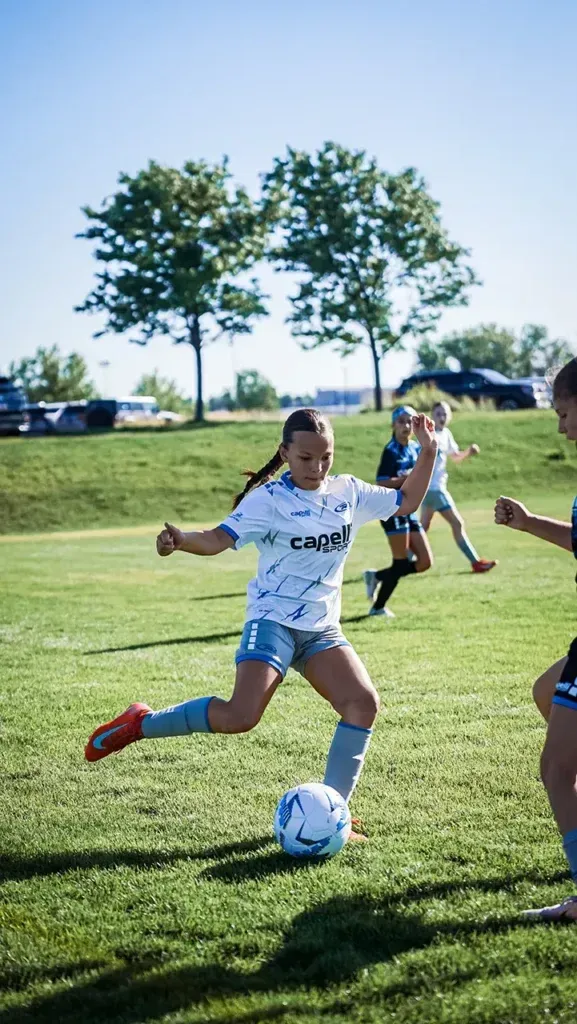 Young girl kicking a soccer ball on a green field under a sunny sky. Other players in the background.