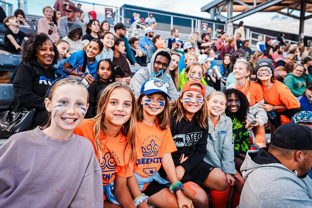 People in bleachers, some with face paint, smiling and looking at the camera at an outdoor event.