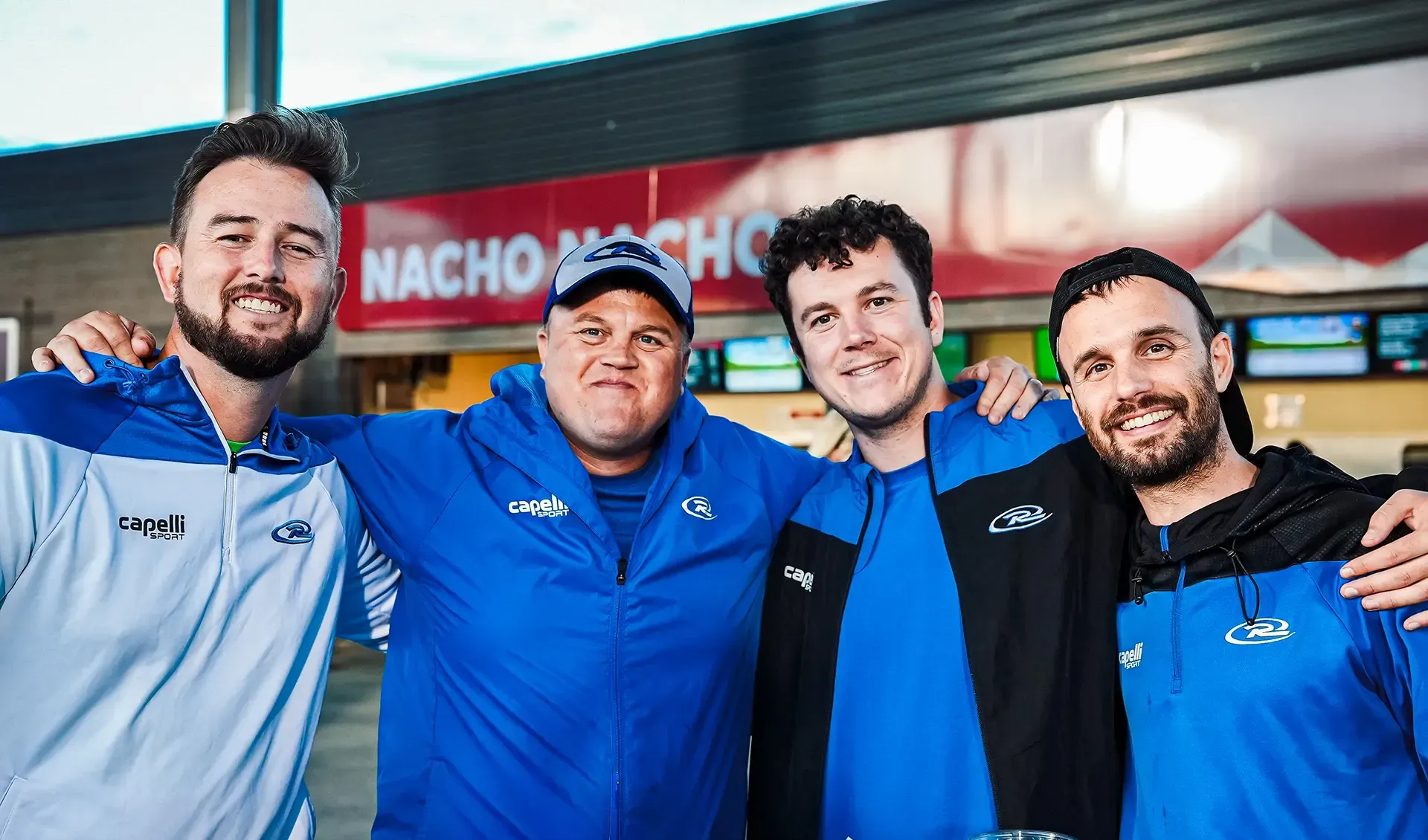 Three people smiling, sitting outdoors: one woman in sunglasses, a man in a white hat, and a man in a grey jacket.