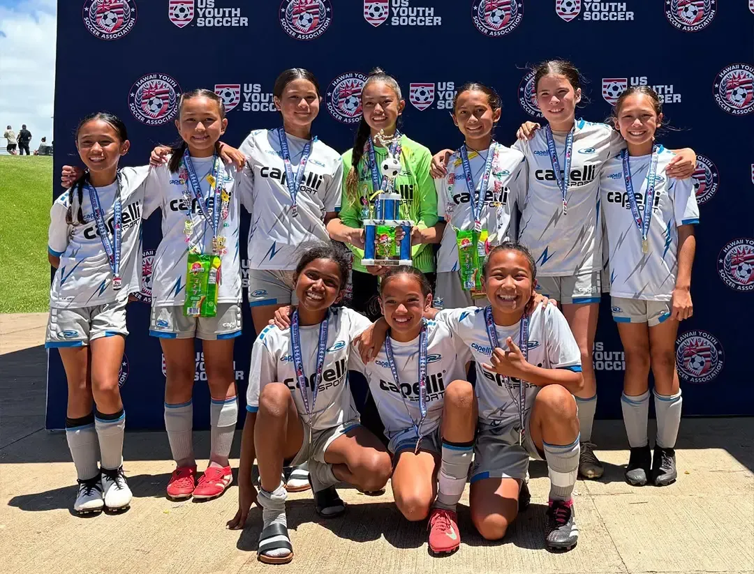 Soccer team poses outdoors, holding trophy and medals. Players wear white and gray uniforms with blue trim.