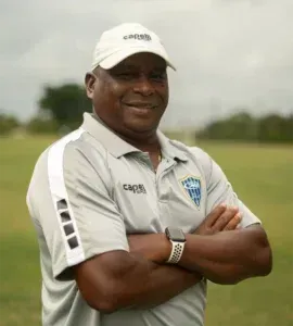 Man wearing cap and polo with arms crossed, standing on a field.