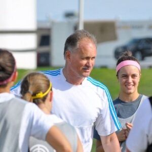 A coach in a white shirt with blue stripes talking to a group of soccer players on a field.