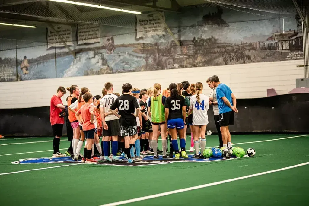 Youth soccer team huddle on indoor field; players in various colored jerseys listen to a coach.