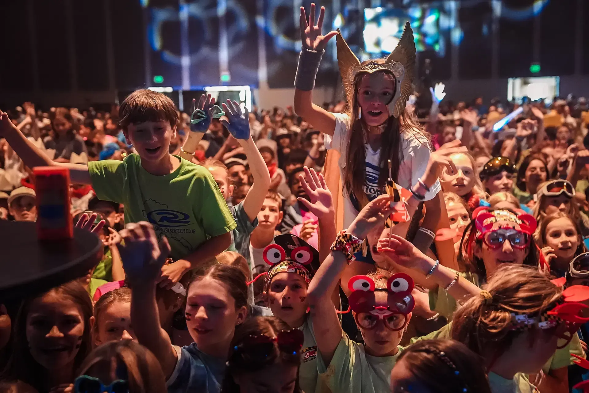 Children waving and smiling at an event with lights. A person is on another's shoulders.