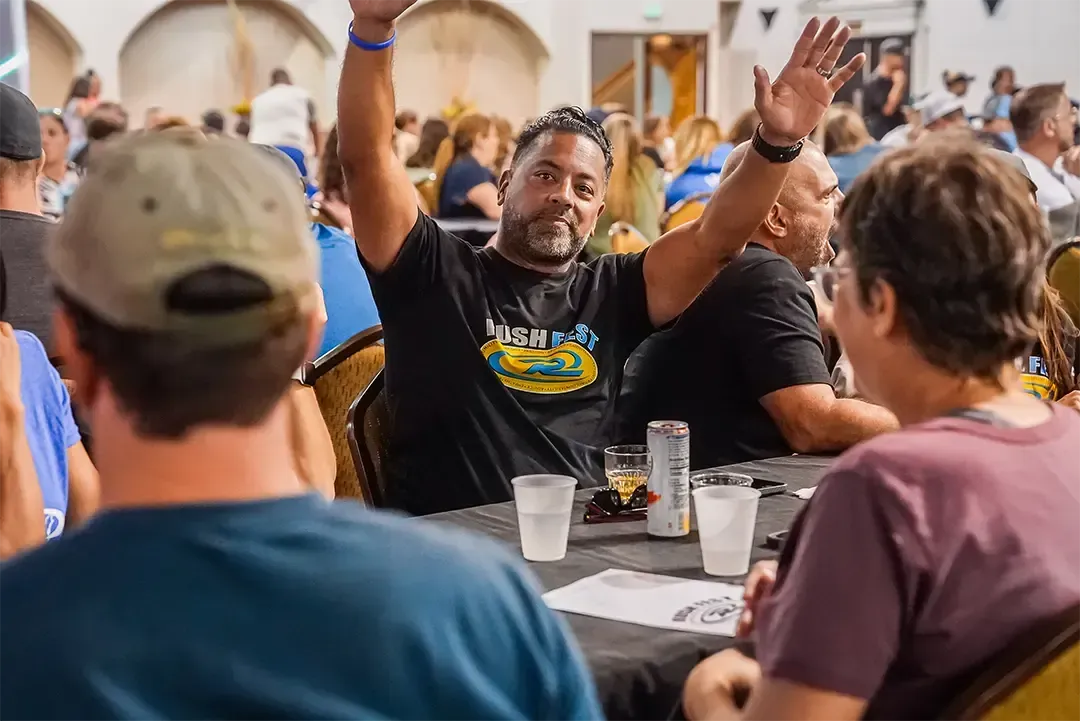 Man with raised arms, wearing a black shirt, at a table in a crowded indoor event, gesturing with excitement.