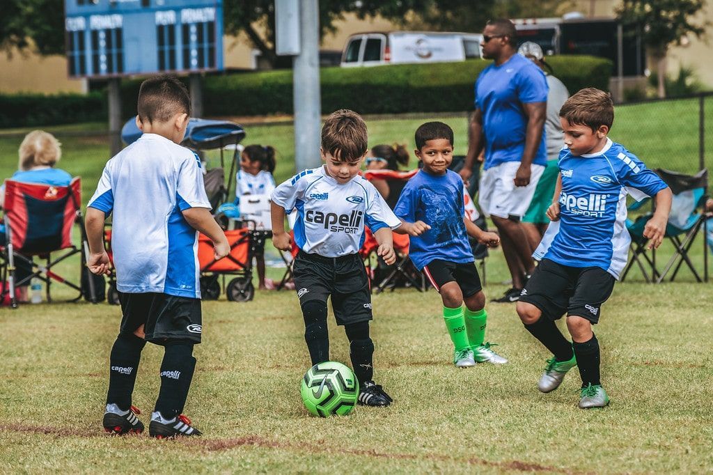 Children playing soccer on a grassy field; blue and white uniforms, adult coaching in the background.