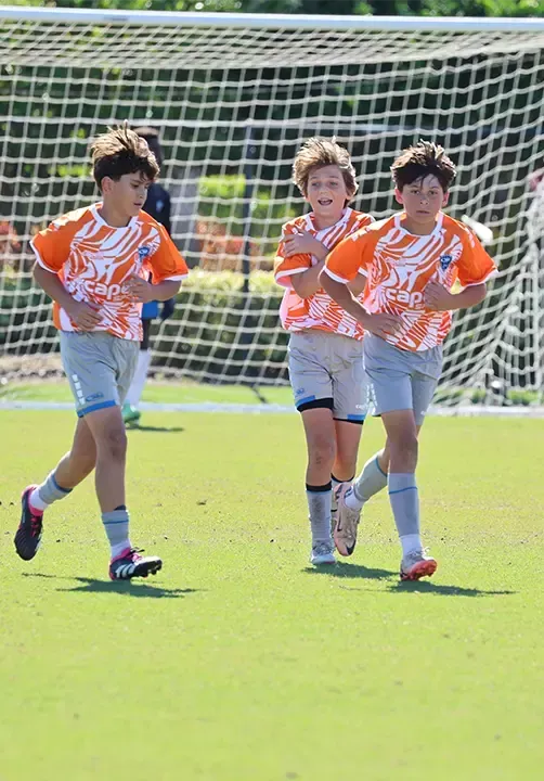 Three soccer players in orange jerseys, running on a green field. A goal net is in the background.