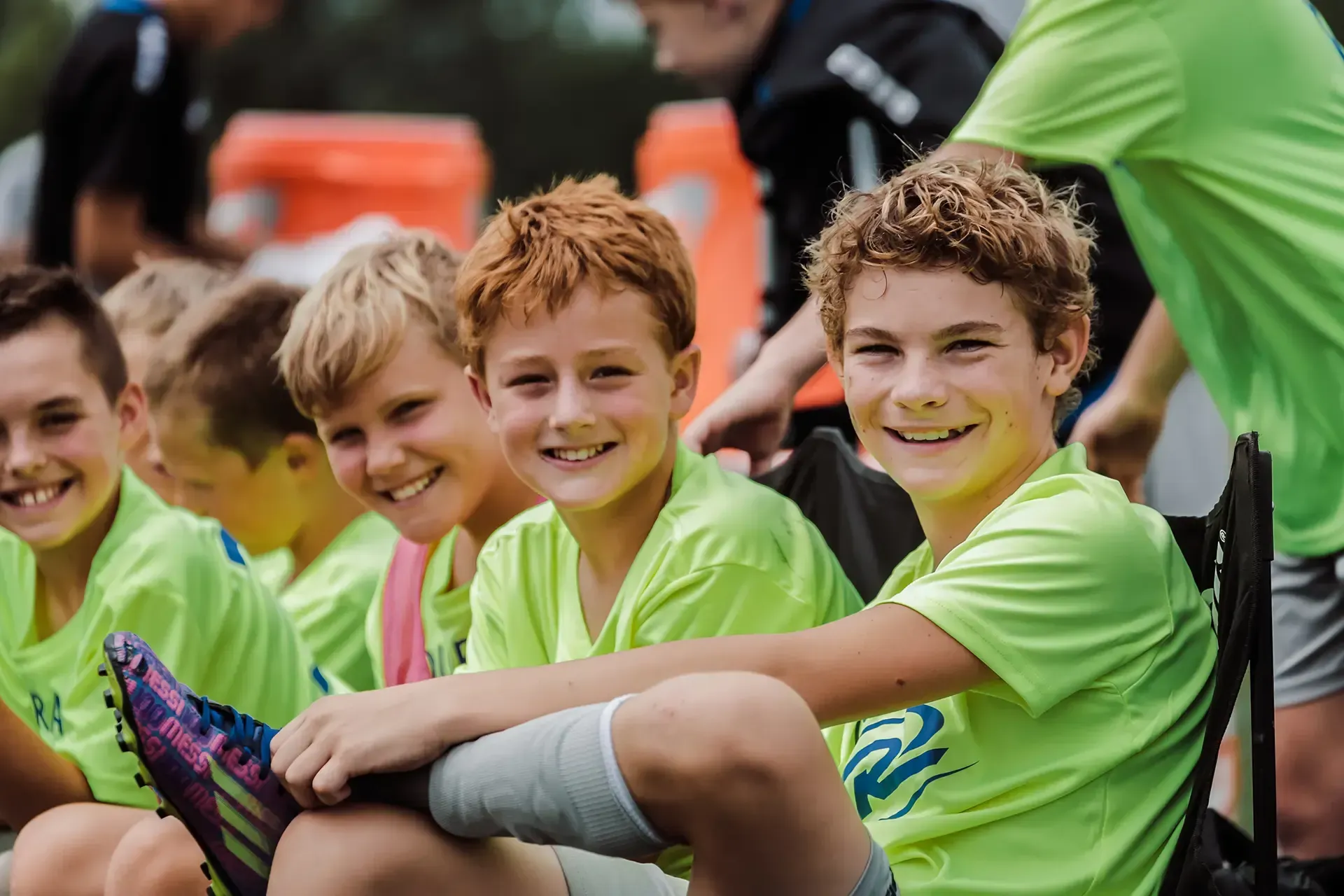 Boys in neon green shirts smiling on a bench at a soccer game.