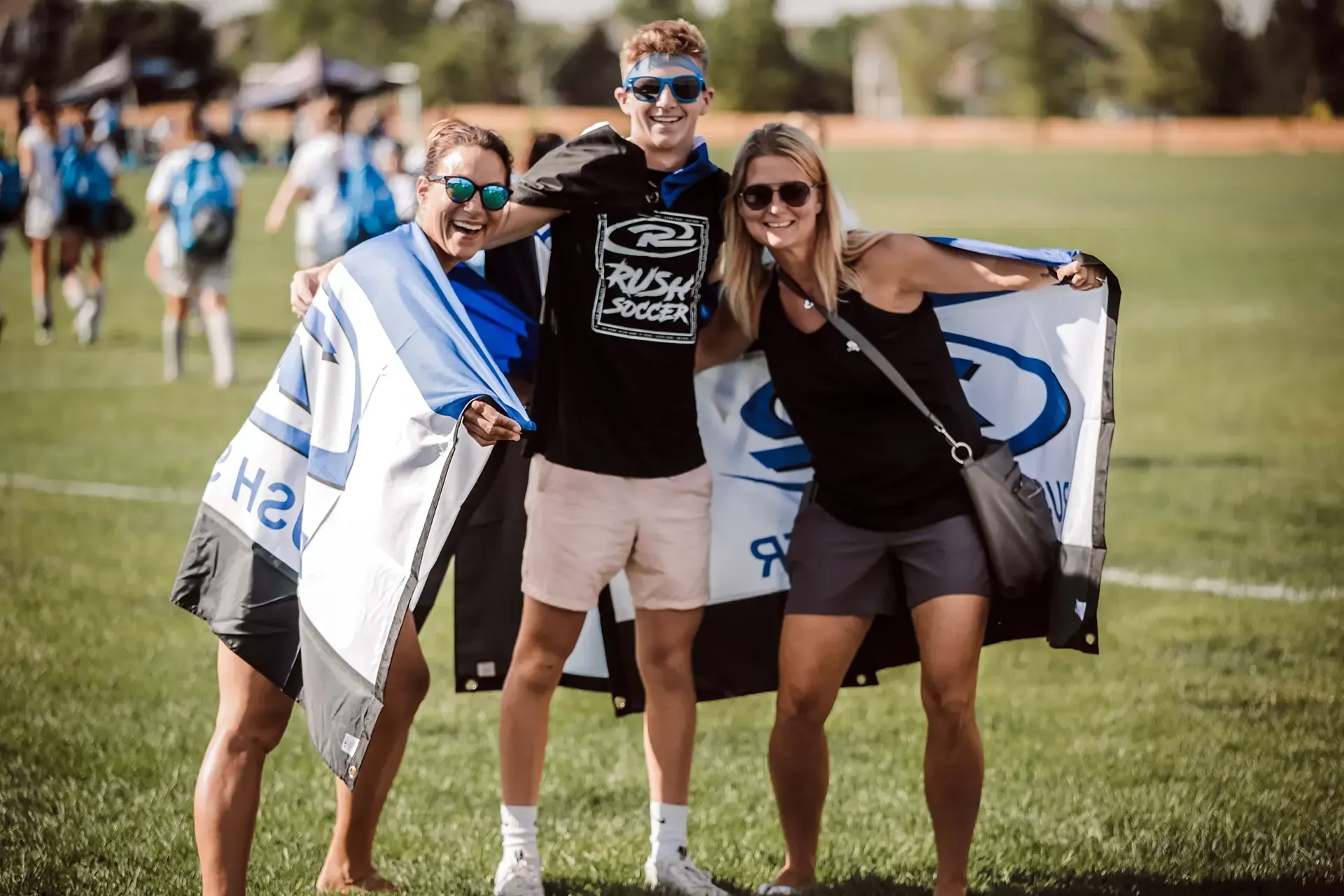 Three people smiling, holding flags with logos on a grassy field; sunny day.