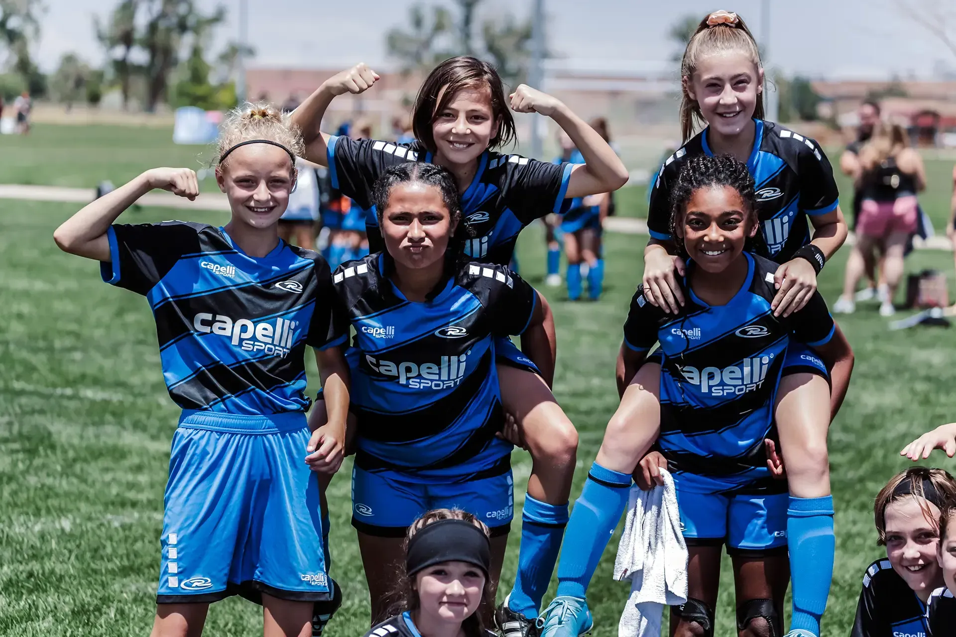 Soccer team celebrates on a field. Girls wear blue and black jerseys, flexing muscles and smiling.
