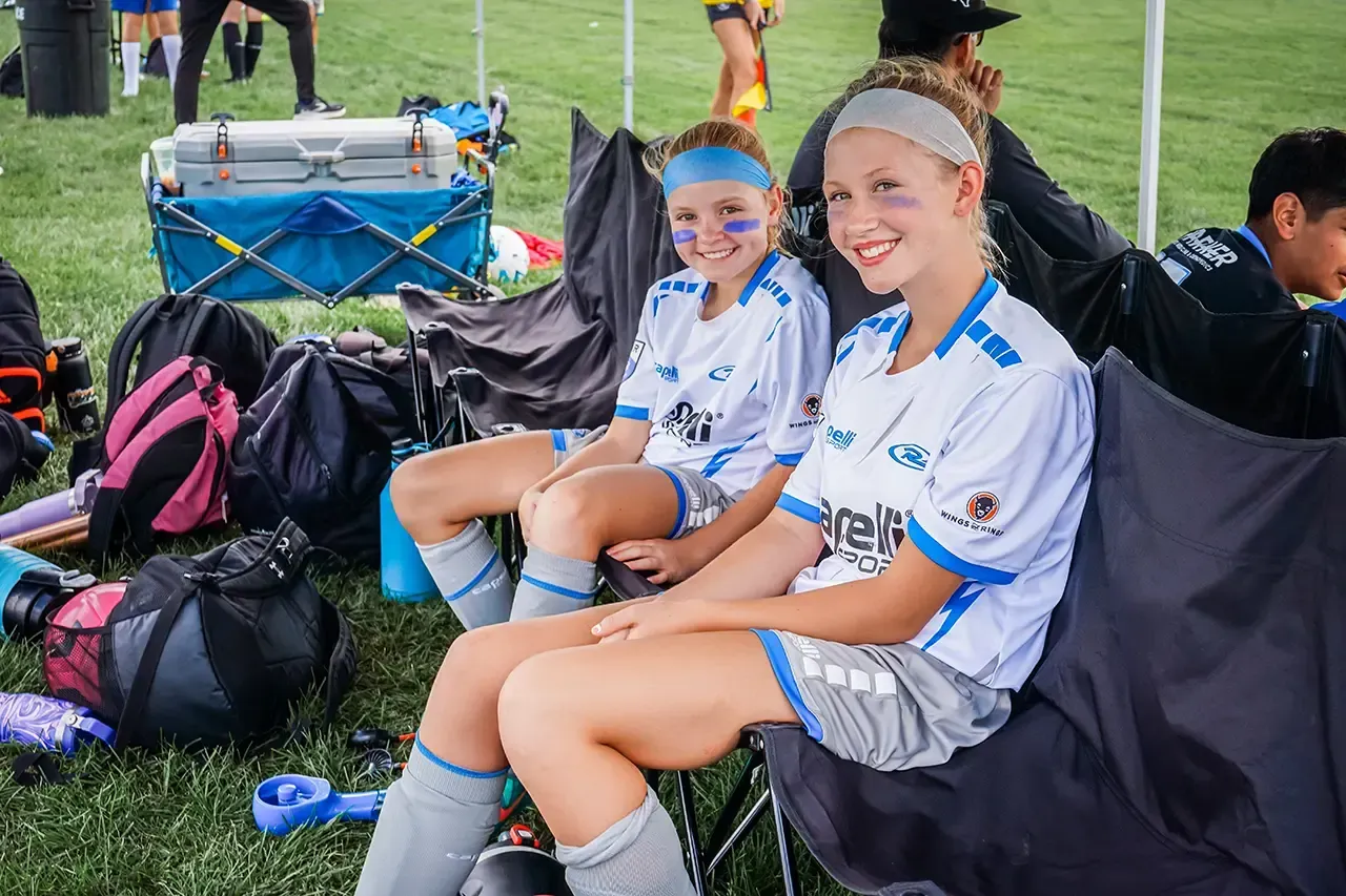 Two soccer players in white jerseys with blue accents sit on folding chairs under a tent, smiling.