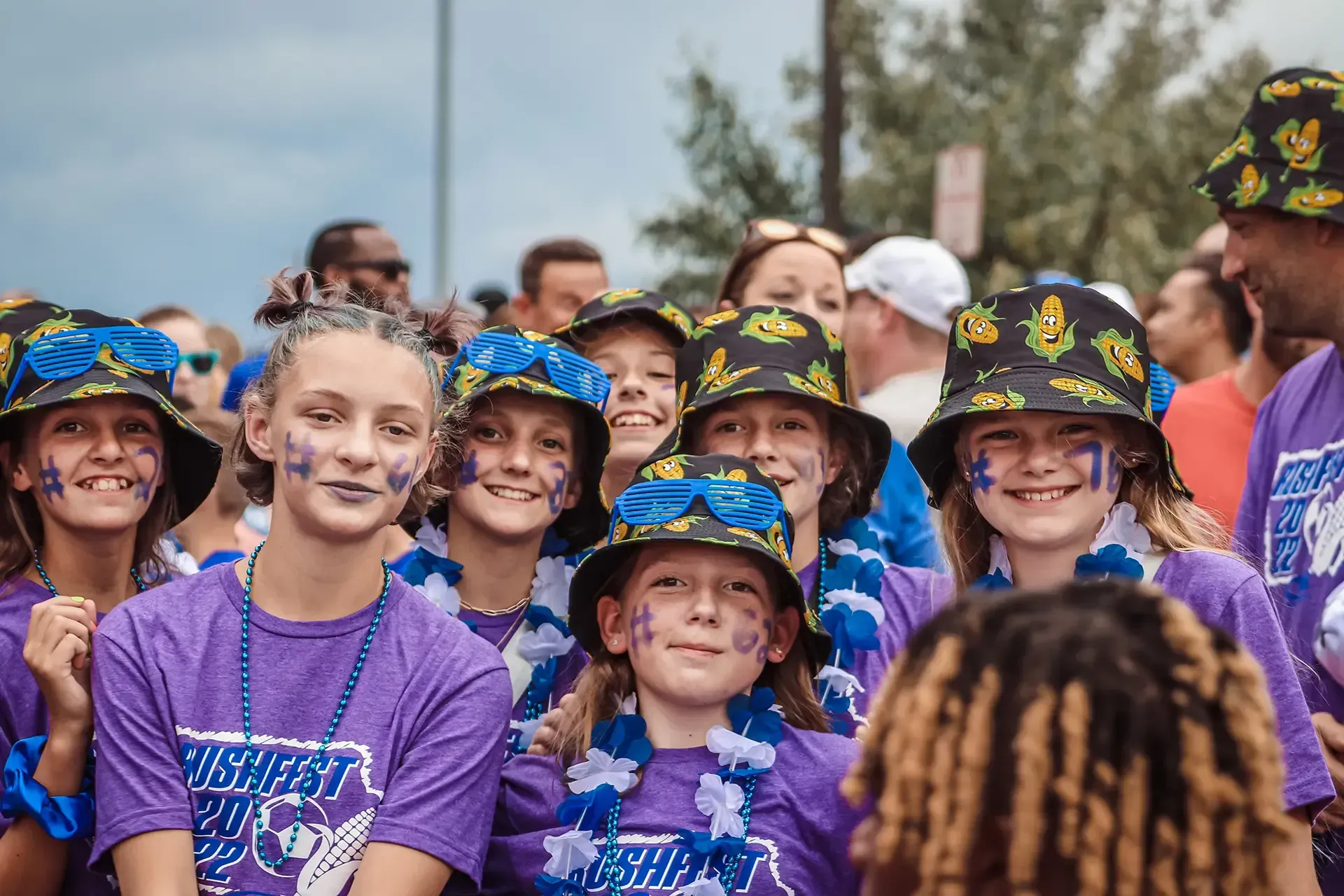 Group of smiling people wearing purple shirts, blue leis, and face paint, celebrating outdoors.