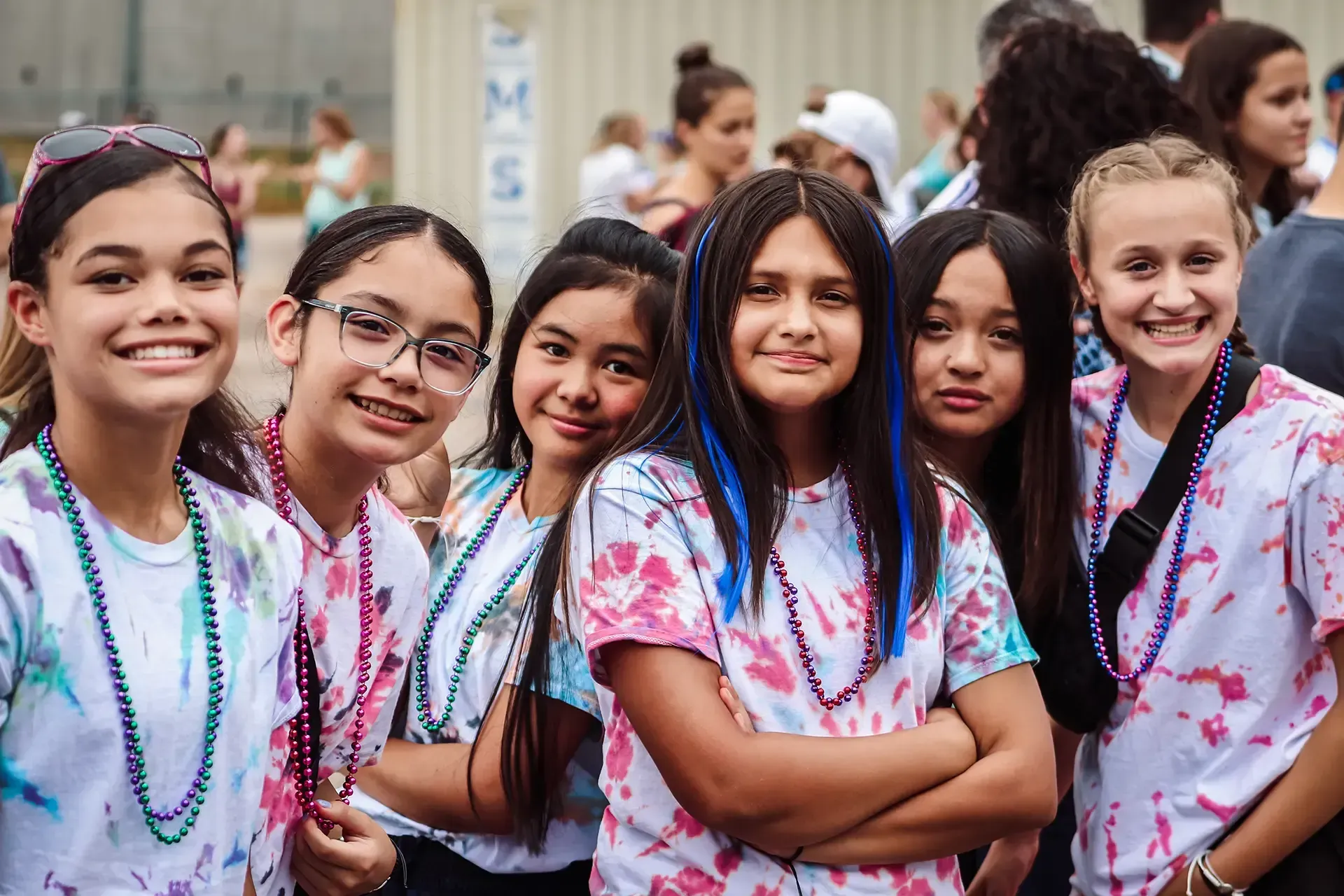 Group of smiling girls in tie-dye shirts and beaded necklaces outdoors.
