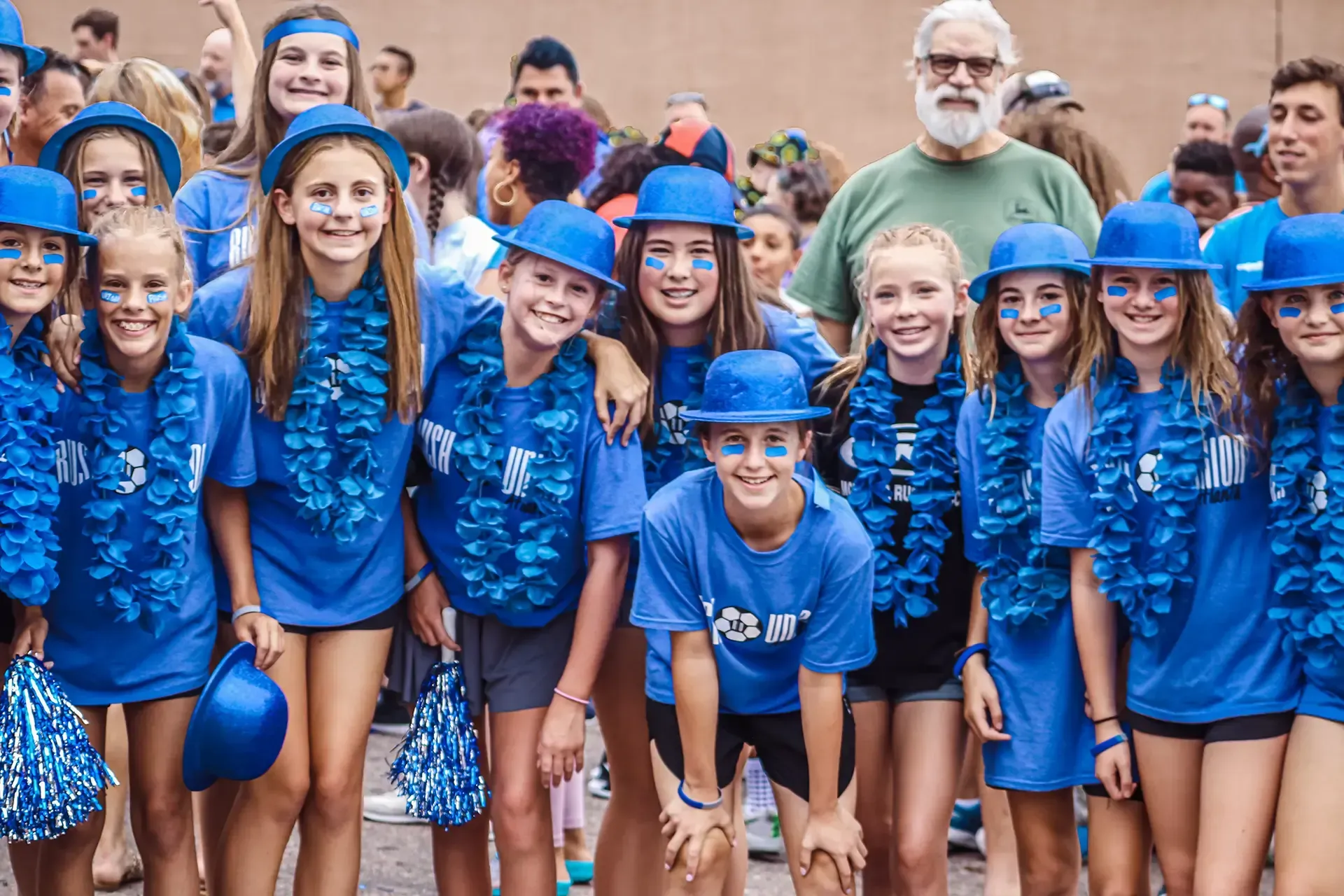 Group of people wearing blue shirts, hats, and leis smiling at the camera, posing outdoors.