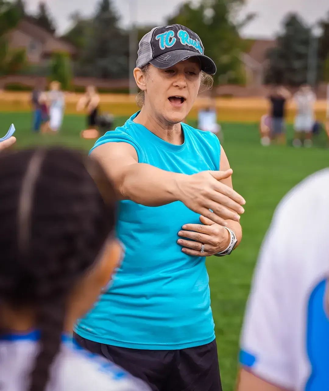 Woman in blue top coaches athletes on a green field, gesturing with her arm.
