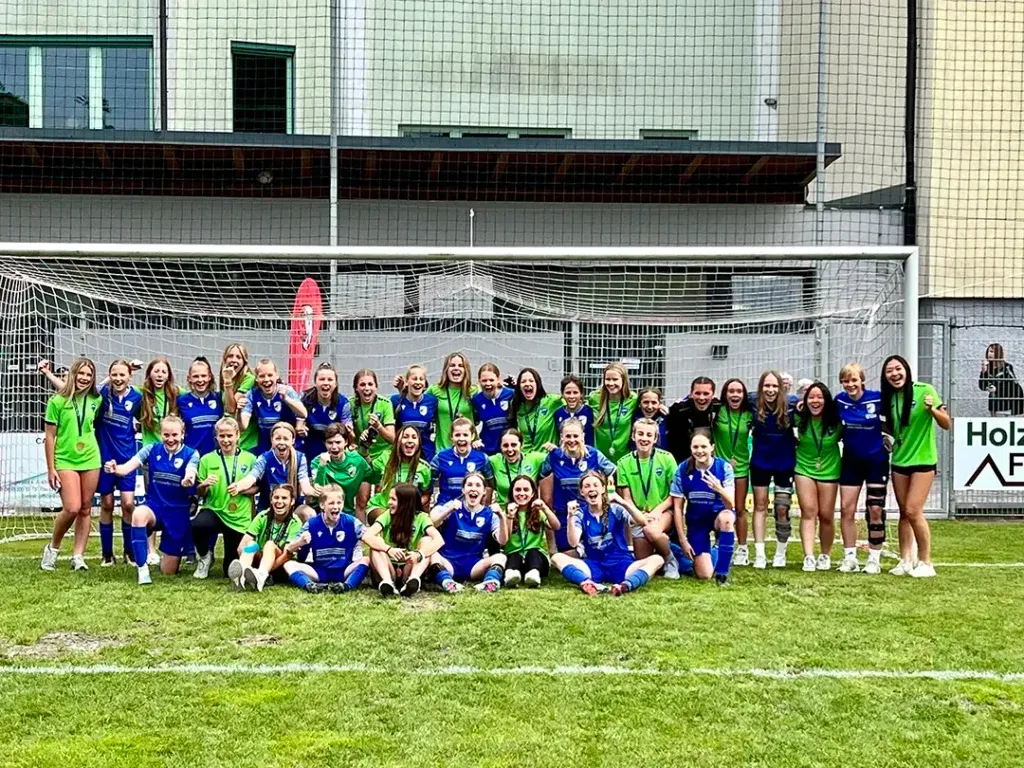 Soccer team poses on a field in front of a goal, many wearing blue and green jerseys.