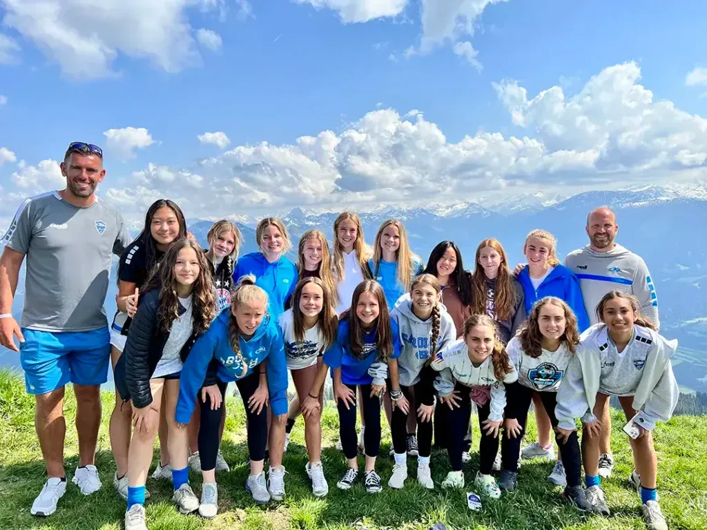 Group of people posing outdoors on a hilltop with mountains in the background. Many wear blue.
