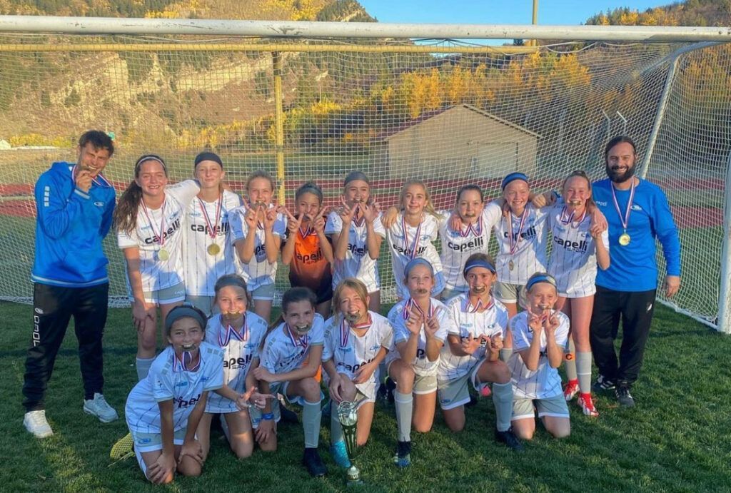 Youth soccer team posing with medals in front of goal, mountains in background.